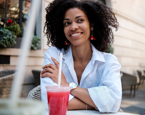 Happy black woman sitting on terrace table with a strawberry juice cup. It has a straw. She is looking away. Summer day.
