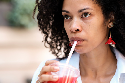Head-shot of curly black woman drinking strawberry juice outdoors. She is drinking from a plastic cup with a straw. She is looking away. No-alcohol beverages. Health lifestyle concept.