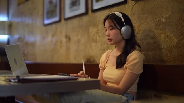 Asian woman working on her laptop and jotting down notes in a cafe, enjoying the flexibility of remote work. Creating a stress-free workspace for enhanced productivity