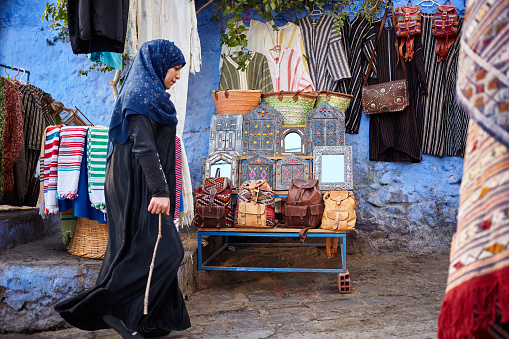 Shopping street with souvenirs shop and woman walking in traditional clothing side view in the blue city of Chefchaouen, Morocco, North Africa.