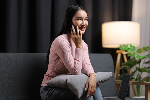 Young Asian woman sitting on sofa at home using mobile phone to send text messages and talk on the phone with her friends.