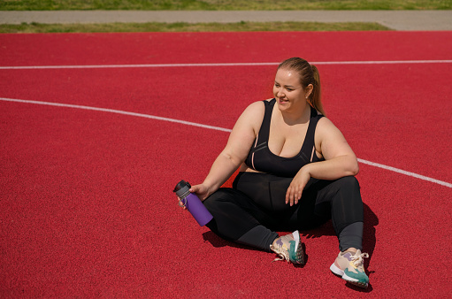 A smiling, cheerful, full-bodied woman with a bottle of water sits on the stadium floor. Relaxation and thirst quenching after sports training