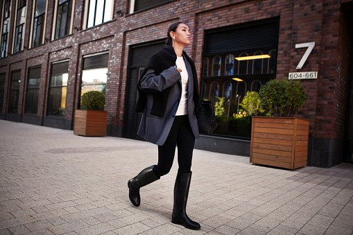 Stylish fashion woman strolls down the street in an oversized blazer, black leggings, and knee high rubber boots. Fashionable outfit, street style.