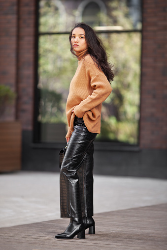 Beautiful Asian woman fashion model in the latest trends layered knit sweater, leather pants, high heeled boots Standing on a city street.