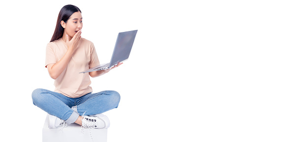 Excited young asian woman sit on white chair with legs crossed and using laptop computer on light pink background copy space Full body young smiling happy female wearing t shirt and jeans lifestyle