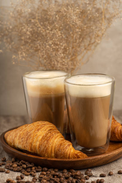 Two lattes with foam tops beside a golden croissant on a wooden tray, surrounded by coffee beans stock photo