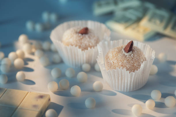 Two coconut truffles with almonds atop, amidst white chocolate chips and bar pieces on a light blue surface