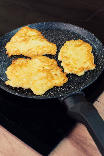 Golden-brown potato pancakes frying in a speckled non-stick pan on a hob. stock photo