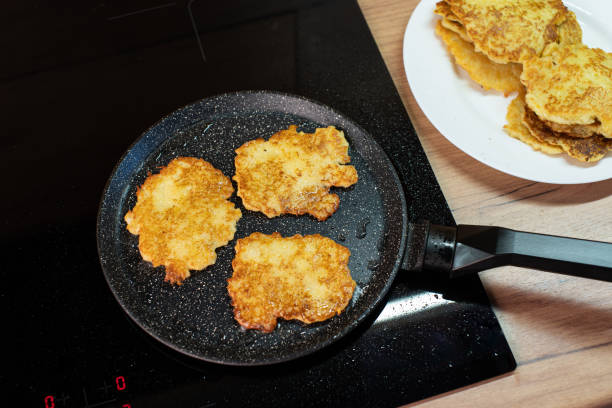 Golden-brown potato pancakes frying in a speckled non-stick pan on a hob. stock photo