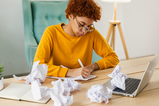 Serious African woman with crumpled paper having problem writing by hand document studying learning at home. Girl no idea sitting at workplace writing notes at office. Start up business deadline