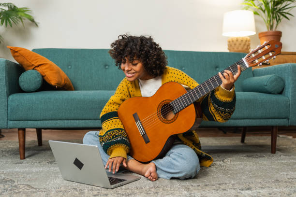 istock African American woman learning guitar watching tutorial on laptop at home. Black girl playing guitar singing song learning online music lessons, Artistic woman playing acoustic guitar learning chords 2073598366