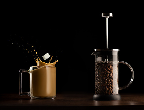 Sugar cube splashing into coffee mug, French press with coffee beans beside, on dark surface