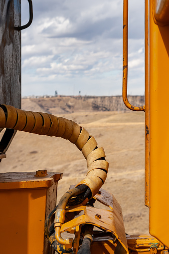 Close-up of the areas between the back of the cabin, and front of the bucket a large yellow John Deere construction vehicle.  Between these are cables and wires wrapped in a yellow protective cover.  The Lethbridge Coulees, Old Mining buildings and High Level Bridge are in the background.