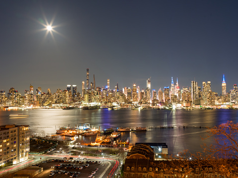 On a full moon night, long exposure image captured using medium format camera system. Manhattan skyline from New Jersey reveals iconic skyscrapers silhouetted against a moody sky, with glittering lights reflecting on Hudson River.