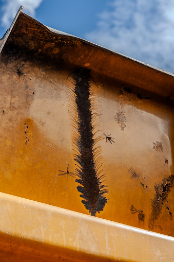 Abstract background - close-up of an orange piece of metal showing dents and rust, including a large long, fringed area of damage and rust.  There is an overhanging piece of metal, Blue sky and white clouds at top of photo