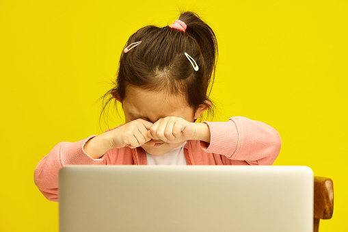 Kid Asian girl in her early years of primary education, expresses signs of emotional distress from the demands of distance learning, wipes tears away or rubbing eyes from fatigue while seated in front of her laptop on a vivid yellow isolated background, encapsulating the unique pressures faced by first graders in a digital learning environment.
