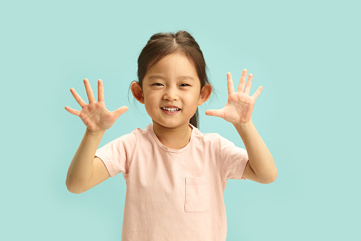 Vivacious Japanese girl, in the age range of 5-7, spreads her hands wide in a high-five motion, her face alight with a jubilant grin, set against a calm blue isolated backdrop, embodying the infectious spirit of childhood cheer