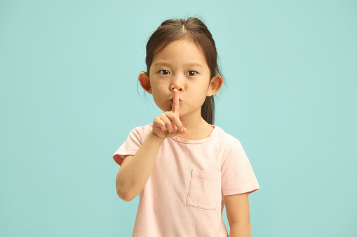 Serious little cute kid asking for silence and quiet, gesturing with finger in front of mouth, saying shh or keeping a secret standing against blue isolated background with a free copy space.