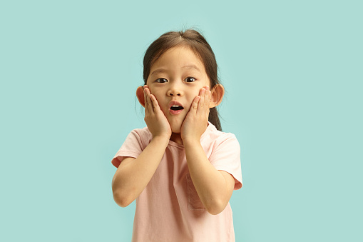 Horizontal photo of Asian ethnicity child with hands on cheeks showing a surprised expression, has amaze face what she saw, found pleasant surprise or received the desired gift, standing against blue isolated background