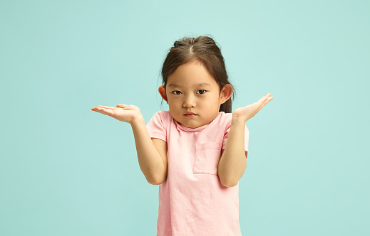 Studio portrait of confused preschool Chinese child girl with shrugged shoulders saying I don't know, questioningly and puzzled, she raised her hands up, showing uncertainty or lack of knowledge standing against blue isolated background with a free copy space. Kid having questionally expression.
