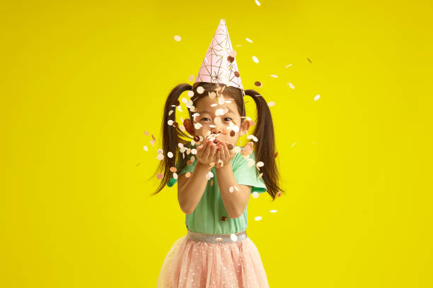 istock Joyful Child with Conical Hat Blowing Confetti from hands, Celebrating Birthday Standing on Bright Yellow Background 2073798556