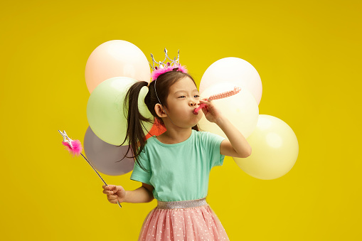 Joyful Chinese little girl in a princess tiara with colorful balloons blowing party whistle on yellow isolated background. Childhood party fun.