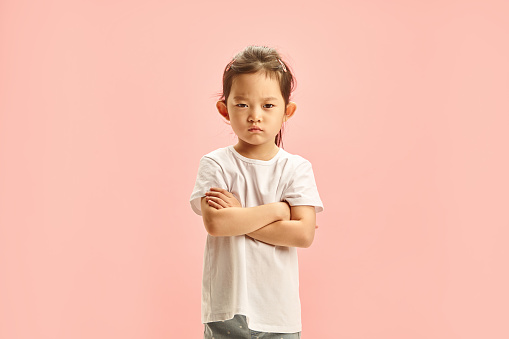 Frontal studio isolated portrait of capricious child kid wearing in white neutral t shirt and jeans, defiantly crossing arms, having dissatisfied and resentment, acting moody, expression frustrated mood standing on a soft pink isolated background with a free copy space. Concept of negativity and children displeased. Younger age crisis.