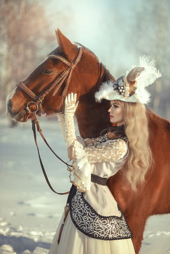 Portrait of a blonde woman with a horse in a winter forest in historical costume. The image of a noble princess. Royal hunt. Artistic photo