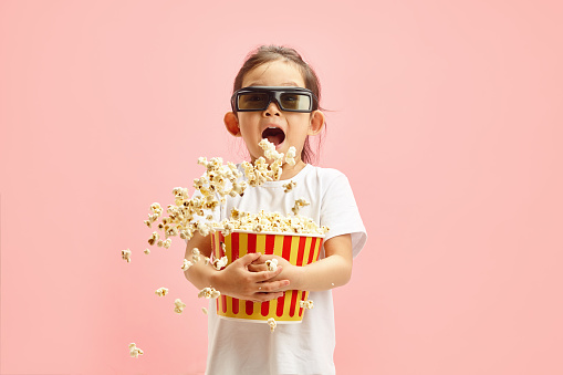Studio portrait of happy little female kid wears in 3D glasses, holding red bucket of popcorn flying to different directions, having an expression of anticipation before the start of cartoon or movie session for bargain price, standing over pink isolated background with a free copy space for your text. Promo action for children movies or visits with kids in morning time in cinemas.
