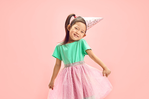 Isolated indoor portrait of five year old Japanese girl wearing in festive clothes, having conical paper hat on head, cute raising shoulders, funny straightening her tutu skirt to the sides, lovely looking at camera standing against soft pink isolated background with a free copy space.