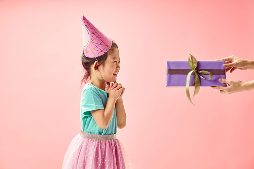 Horizontal shot of hopeful little girl in party hat stands near presented to her gift box, squeeze hands in front of herself believes in good luck makes wish on her birthday wears turquoise t shirt and pink tutu skirt poses over pink isolated