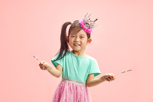 Photo of birthday celebrating little Chinese appearance child girl holding sparklers, wearing in festive clothes and crown princess on head, having cute ponytail, happy smiling looking to camera, having fun poses against soft pink isolated background with a free copy space. Fulfillment of children's desires concept.