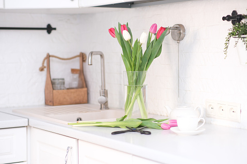 Home kitchen interior.Vase with bouquet of pink and white tulips, glass of tea, sissors, teapot on white worktop of kitchen cupboard. Spring consept. High quality photo