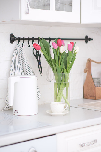 Home kitchen interior.Vase with bouquet of pink and white tulips, glass of tea, electric teapot on white worktop of kitchen cupboard. Spring consept. High quality photo
