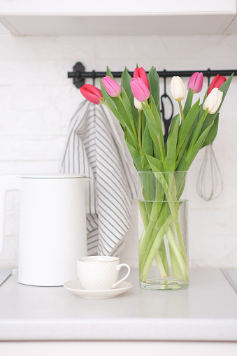 Home kitchen interior.Vase with bouquet of pink and white tulips, glass of tea, electric teapot on white worktop of kitchen cupboard. Spring consept. High quality photo