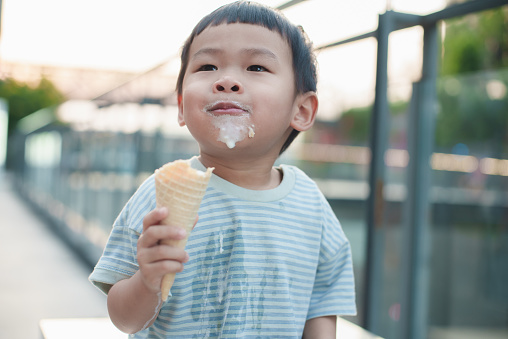 A cute Asian boy with short black hair holding a milk ice cream cone has smears of ice cream on his shirt, cheeks and chin showing his enthusiasm and the messy joy of the moment. An Asian boy happily and messily eating ice cream with a funny expression.