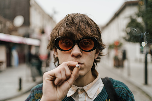 Fashionable young man in retro 90s style attire smokes cigarette on bustling city street, exuding vintage urban vibe.