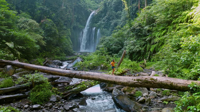 Aerial view of woman crossing river by log on the background of tropical waterfall while hiking