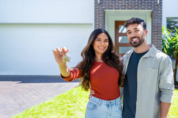 happy woman and man, holding keys from new first house, young family celebrating moving day, happy woman and man, holding keys from new first house, young family celebrating moving day, satisfied customers couple purchase real estate, mortgage and relocation concept. The building can be seen in the background happy family with key of their new house stock pictures, royalty-free photos & images