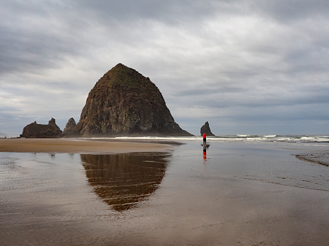 Cannon Beach, Oregon - 9/30/2018: Woman in red windbreaker photographs Haystack Rock and its reflection at extreme low tide under dramatic autumn cloudscape.