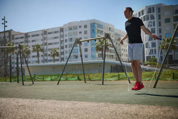 Young athletic man exercising with a jumping rope on the outdoor urban sportsground. Full length portrait of a sportsman, Latin American guy in active wear warming up, jumping through a skipping rope