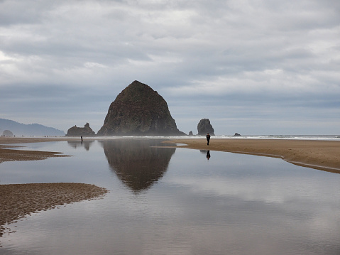 Cannon Beach, Oregon - 9/30/2018: Man photographs Haystack Rock and its reflection at extreme low tide under dramatic autumn cloudscape.