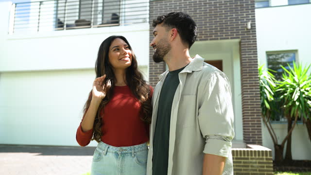 Couple standing proudly in front of their new home. They are both wearing casual clothes and embracing