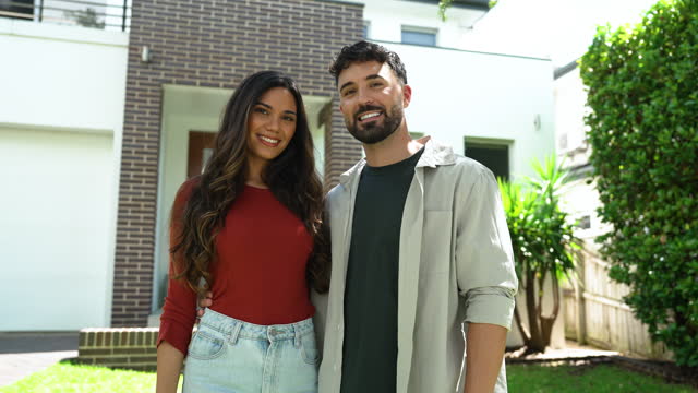Couple standing proudly in front of their new home. They are both wearing casual clothes and embracing