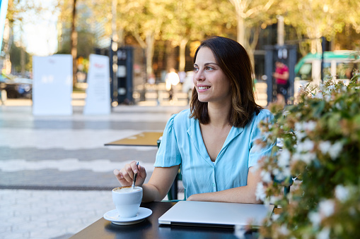 Cheerful young woman in a light blue shirt smiling and stirring her coffee, sitting at an outdoor cafe table with a closed laptop, amidst a lively city backdrop. Concept of distance and remote working