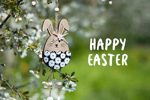 Greeting card with text Happy Easter in English. Easter decor. Decorative wooden Easter bunny toy on background of blooming spring garden. Outdoor