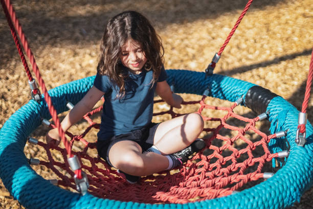 Portrait of a beautiful caucasian girl sits on a round rope swing in the park at the playground Portrait of a beautiful caucasian girl with short brown flowing hair, smiling and looking down with half-closed eyes, sits on a round swing in the park at the playground, close-up side view. The concept of PARKS and REC, happy childhood, children picnic, holidays, children recreation, outdoor recreation, playgrounds, outdoor recreation, family vacation funny camping signs pictures stock pictures, royalty-free photos & images