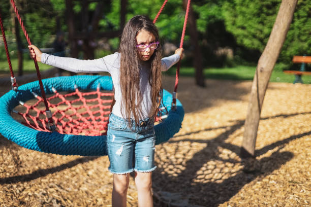 Portrait of a beautiful Caucasian dissatisfied girl with a round rope swing in the park Portrait of one beautiful Caucasian brunette girl of eight years old stands displeasedly holding on to a round rope swing in a park on a playground, close-up side view. The concept of PARKS and RECREATION, happy childhood, children's picnic, children's recreation, outdoor recreation, playgrounds, outdoor recreation, family vacation. funny camping signs pictures stock pictures, royalty-free photos & images