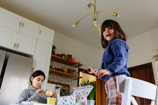 low angle view of two girls at table, one painting the egg while smaller girl stands on chair smiling