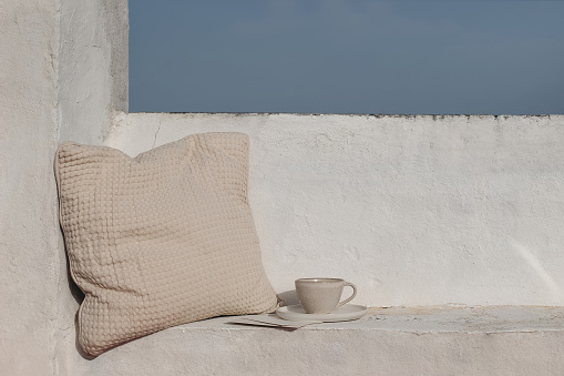 Summer vacation still life, relaxation concept. Mediterranean outdoor sitting area. Bench with beige waffle textured cushion. Cup of coffee. White wall background. Blue sky. Travel banner